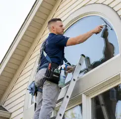 Professional window cleaner on a ladder using a squeegee to clean an exterior arched window, Yonkers, NY.